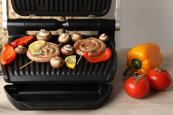 Electric grill with homemade sausages, bell peppers and mushrooms on wooden table