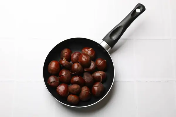 stock image Fresh edible sweet chestnuts in frying pan on white tiled table, top view