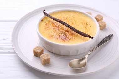 Lezzetli creme brulee kasesi, vanilya kapsülü, şeker küpleri ve kaşık beyaz ahşap masa, yakın çekim