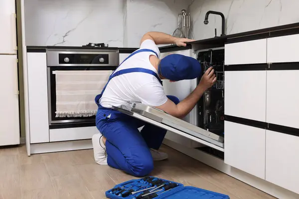 Serviceman repairing dishwasher with screwdriver in kitchen