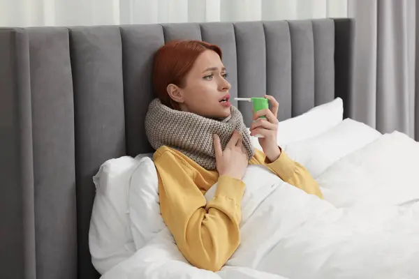 Young woman with scarf using throat spray in bed