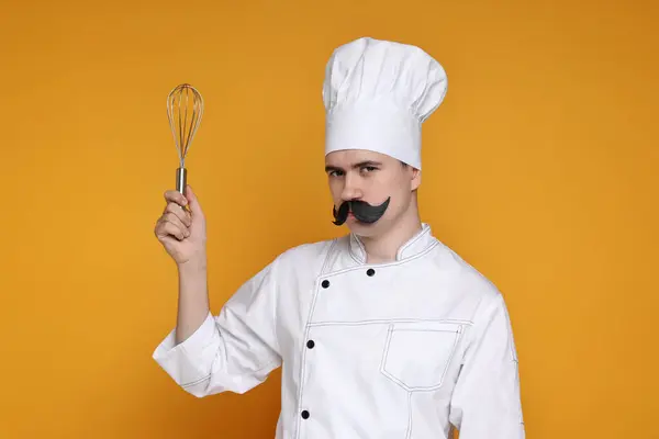 Portrait of happy confectioner with funny artificial moustache holding whisk on orange background