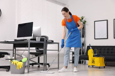 Temizlik işçisi yerleri paspasla yıkıyor. Ofiste içi malzeme dolu kova ve ıslak tabela var.