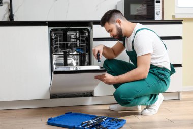 Görevli mutfakta tornavidayla bulaşık makinesinin kapısını tamir ediyor.