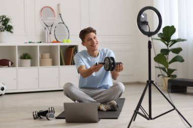 Gülümseyen spor blogcusu evde akıllı telefonuyla çevrimiçi fitness dersini izlerken kas silindiri tutuyor.