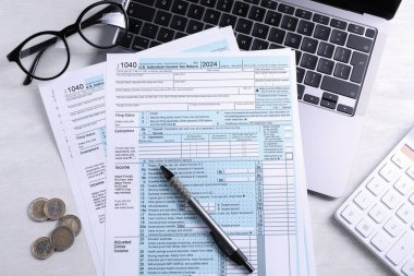Açık gri masa üzerinde vergi formları, madeni paralar, kalem, dizüstü bilgisayar ve hesap makinesi, düz konum