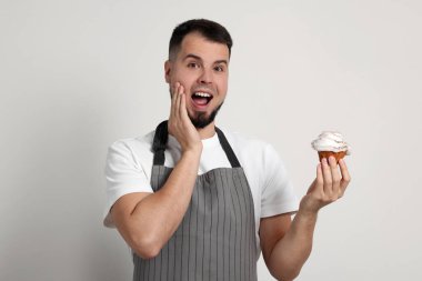 Önlüklü mutlu profesyonel pastacı açık gri arka planda lezzetli kekler tutuyor.