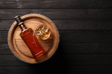 Whiskey with ice cubes in glass, bottle and barrel on black wooden table, top view. Space for text clipart