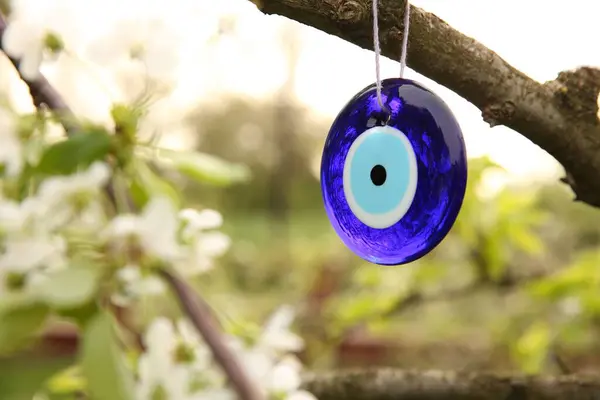 Evil eye amulet hanging on blossoming spring tree outdoors, closeup. Space for text