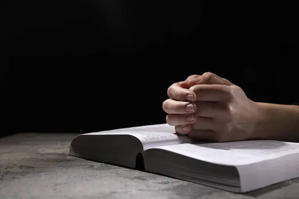 Stock image Religion. Christian woman praying over Bible at table against black background, closeup. Space for text