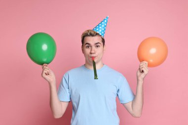 Parti şapkalı genç adam, pembe arka planda balonlar ve üfleyici.