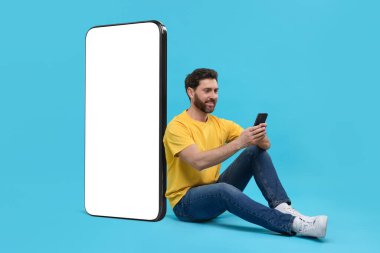 Cep telefonu olan bir adam açık mavi arka planda boş ekranlı büyük bir cihazın yanında oturuyor. Tasarım için model