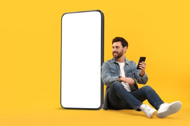 Cep telefonu olan bir adam turuncu arka planda boş ekranı olan büyük bir cihazın yanında oturuyor. Tasarım için model