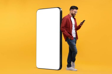 Cep telefonu olan bir adam turuncu arka planda boş ekranlı dev bir cihazın yanında duruyor. Tasarım için model