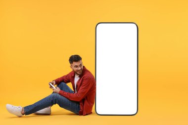 Cep telefonu olan bir adam turuncu arka planda boş ekranı olan büyük bir cihazın yanında oturuyor. Tasarım için model