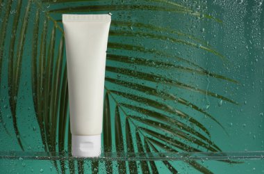 Yeşil zemin üzerinde nemlendirici krem ve palmiye yaprağı olan bir tüp. Islak cam üzerinden görüş.