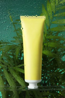 Yeşil zemin üzerinde nemlendirici krem ve palmiye yaprakları olan bir tüp. Islak cam üzerinden görüş.