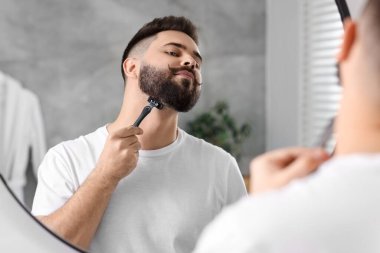 Yakışıklı genç adam banyoda aynanın yanında tıraş oluyor.