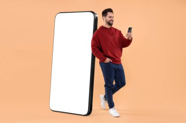 Cep telefonu olan bir adam koyu bej arka planda boş ekranlı büyük bir cihazın yanında duruyor. Tasarım için model