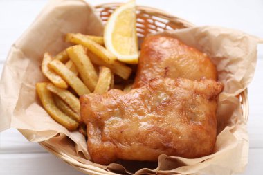 Lezzetli balık, patates kızartması ve limon hasır kasede beyaz ahşap masada, yakın plan
