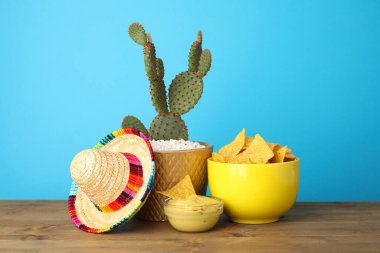 Meksika sombrero şapkası, kaktüs, nachos cipsi ve guacamole kaselerde açık mavi arka planda, ahşap masada.