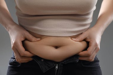 Gri arka planda göbek yağına dokunan kadın, yakın plan. Aşırı kilolu