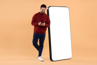Cep telefonu olan bir adam koyu bej arka planda boş ekranlı büyük bir cihazın yanında duruyor. Tasarım için model