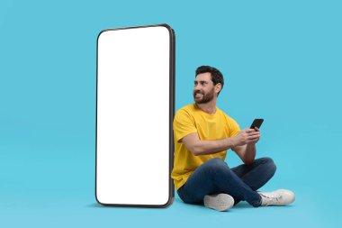 Cep telefonu olan bir adam açık mavi arka planda boş ekranlı büyük bir cihazın yanında oturuyor. Tasarım için model