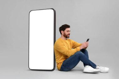 Cep telefonu olan bir adam gri arka planda boş ekranı olan büyük bir cihazın yanında oturuyor. Tasarım için model
