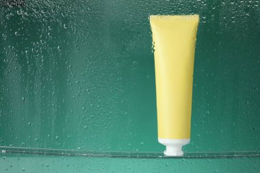 Yeşil arka planda nemlendirici krem bulunan bir tüp. Islak camdan görülüyor. Metin için boşluk