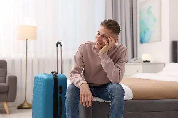 Smiling guest with suitcase relaxing on bed in stylish hotel room
