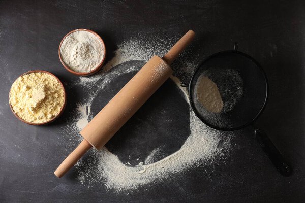 Scattered flour, rolling pin and sieve on black table, flat lay