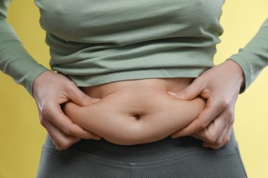 Sarı arka planda göbek yağına dokunan kadın, yakın plan. Aşırı kilolu