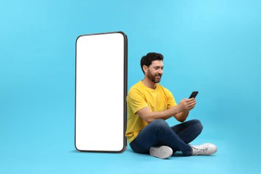 Cep telefonu olan bir adam açık mavi arka planda boş ekranlı büyük bir cihazın yanında oturuyor. Tasarım için model