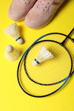 Tüylü badminton mekikleri, raketler ve spor ayakkabıları sarı arka planda, düz yatıyordu