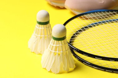 Tüylü badminton mekikleri, raketler ve spor ayakkabıları sarı arka planda, yakın plan