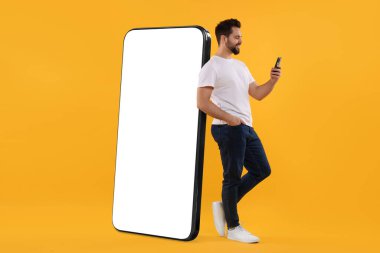 Cep telefonu olan bir adam koyu bej arka planda boş ekranlı büyük bir cihazın yanında duruyor. Tasarım için model