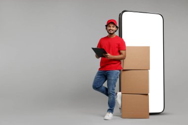Gri arkaplanda kocaman bir akıllı telefonun yanında paket ve pano yığını olan bir kurye. Teslimat servisi. Metin için boşluk