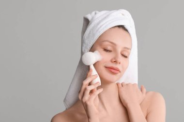 Genç bir kadın gri arka planda yüzünü fırça ve temizleyici köpükle yıkıyor. Metin için boşluk