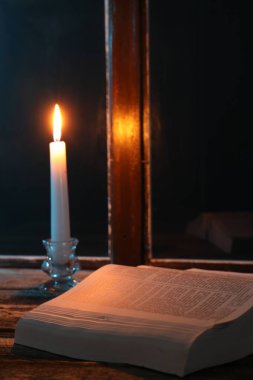 Geceleri pencerenin yanındaki tahta masada yanan mum ve İncil.