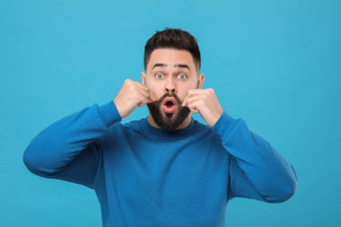 Açık mavi arka planda bıyığa dokunan genç adam şaşırdı.
