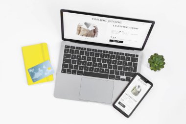 İnternetteki web sitesi laptop ekranında. Bilgisayar, akıllı telefon, not defteri, kredi kartı ve beyaz arka planda ev bitkisi, düz konum
