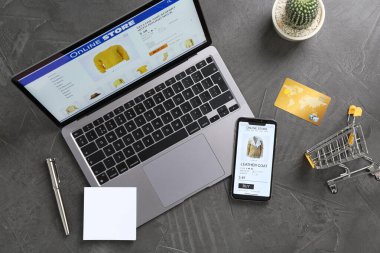 İnternetteki web sitesi laptop ekranında. Bilgisayar, akıllı telefon, kredi kartı, kırtasiye, alışveriş arabası ve kaktüs gri masada, düz yerde.