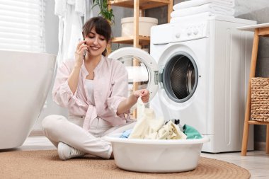 Çamaşırları yıkanmış, evde çamaşır makinesinin yanında akıllı telefondan konuşan mutlu bir ev hanımı.