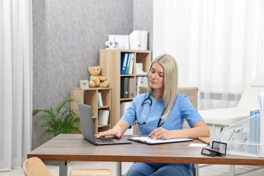 Klinikteki masada dosya ve dizüstü bilgisayarla çalışan bir doktor.