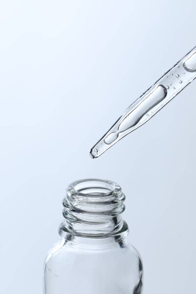 Dripping liquid from pipette into glass bottle on light blue background, closeup