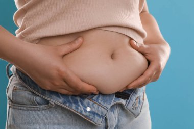 Açık mavi arka planda göbek yağına dokunan kadın, yakın plan. Aşırı kilolu