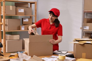 Postane çalışanı içeride ahşap masada paket paketliyor.