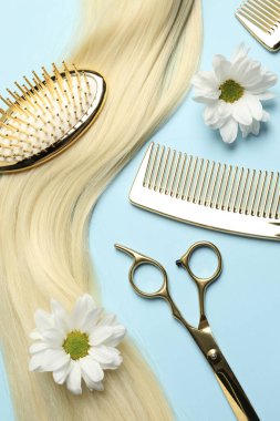 Profesyonel kuaför aletleri, çiçekler ve açık mavi arka planda sarı saç teli olan düz kompozisyon.