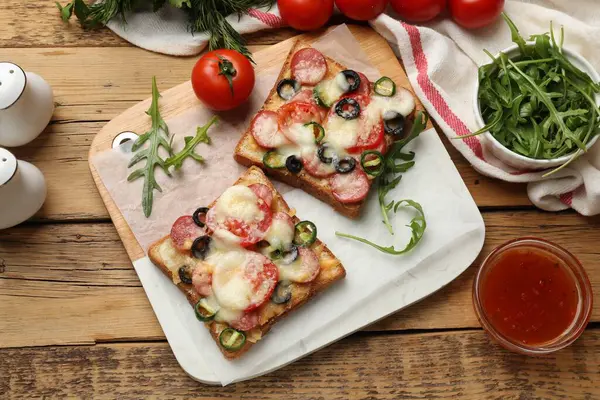 Tasty pizza toasts, sauce, arugula and tomatoes on wooden table, top view